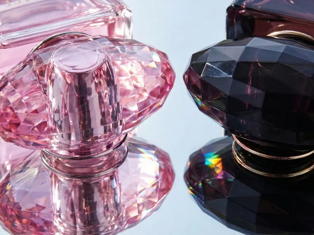 A macro photograph of the crystal stoppers of the Bright Crystal and Crystal Noir perfume bottles. They are lying side-by-side on a mirrored surface. Light passes through the facets of the glass, creating rainbow refractions (a prismatic effect) on the surface. The emphasis is on the weight, transparency, and perfect cut of the glass.