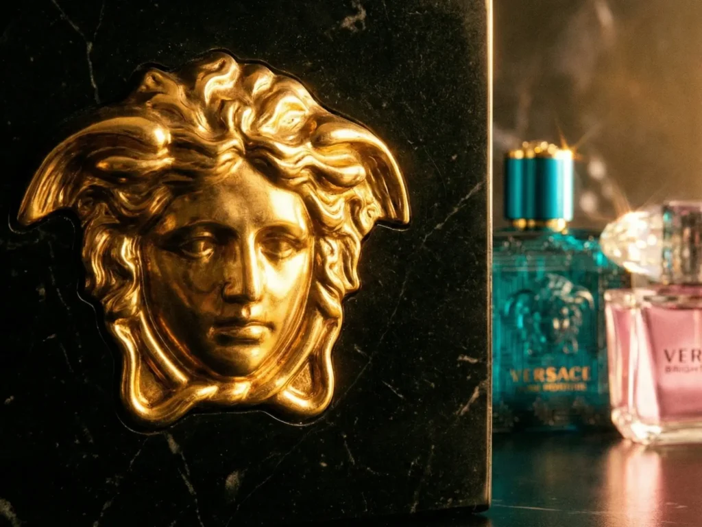 A dramatic, cinematic composition. A detailed close-up photograph of the gold Medusa Gorgon bas-relief logo on a textured, dark surface like polished black marble or rich velvet. Several Versace perfume bottles stand out of focus in the background, including a turquoise Eros and a pink Bright Crystal, catching glints of light. The lighting is warm, golden, and highly contrasting, creating deep shadows and brilliant highlights.